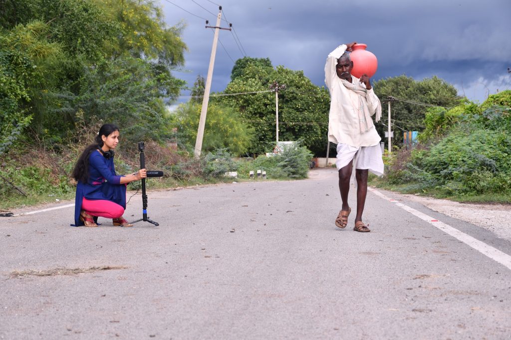 Woman setting up a camera on a road beside a man carrying a red pot on his shoulder